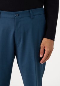 Pantalons à motifs bleu foncé avec une texture lisse, dotés d'une fermeture à bouton et de poches latérales, présentés avec une main positionnée près de la taille.