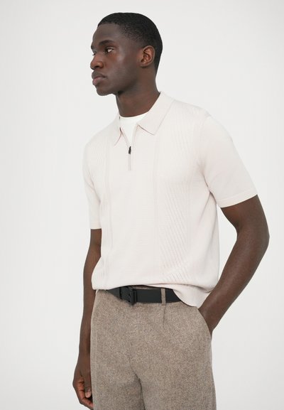 Ανοιχτό ροζ polo κοντομάνικο με φερμουάρ στον γιακά και ριγωτή υφή. Συνδυασμένο με καφέ υφασμένα παντελόνια και μαύρη ζώνη.