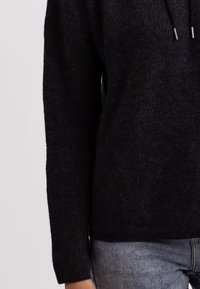 Maglione nero a maglia con maniche lunghe, texture a coste e cappuccio con cordoncino. Abbinato a jeans azzurri, creando un look casual.