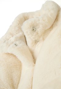 Tejido de piel sintética de color crema con una textura suave, que cuenta con un cuello y cierres de presión plateados en un diseño suavemente curvado.