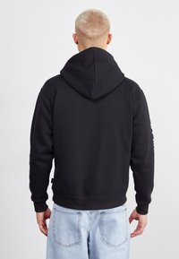 Sudadera negra con una capucha grande, puños acanalados y un cordón ajustable. La tela parece suave con una textura lisa. Jeans de color azul claro visibles.