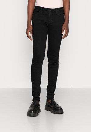 Persoon die strakke zwarte broek en zwarte stevige plateauzolen draagt, staat tegen een effen lichte achtergrond.