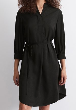 Mujer con un vestido negro hasta la rodilla, mangas tres cuartos, escote en V y un cinturón delgado atado a la cintura, sobre un fondo liso.