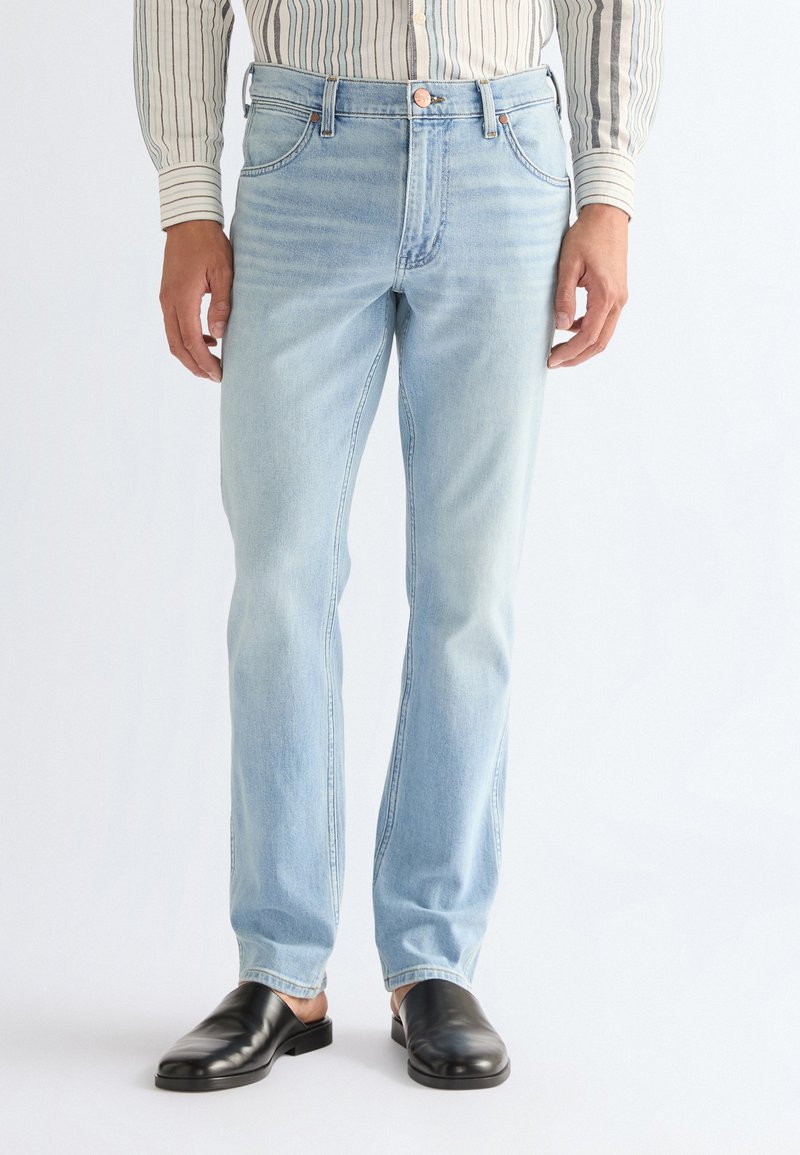 Τζιν ελαφρώς μπλε με ίσια γραμμή που φοριέται με ριγέ πουκάμισο και μαύρα παπούτσια τύπου slip-on σε απλό φόντο.