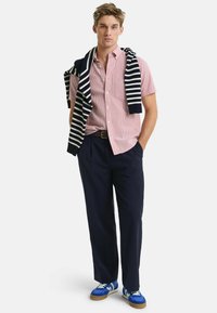 Rot-weißer karierten Kurzarmhemd, marineblaue Hose, marineblauer gestreifter Pullover über die Schultern gelegt, blaue Turnschuhe mit weißen Schnürsenkeln und Gummisohlen.