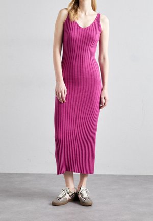 Woman wearing a sleeveless, ribbed, magenta midi dress and checkered sneakers standing against a plain gray wall.