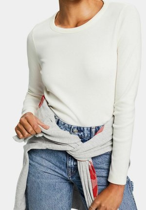 Femme portant un haut blanc à manches longues et un jean bleu avec un sweat-shirt gris noué autour de la taille, montrant une doublure intérieure rouge.