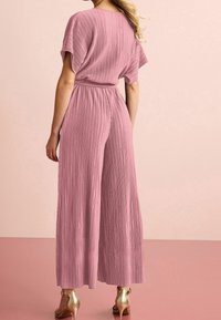 Mujer vestida con un mono plisado de color rosa empolvado con mangas cortas y sandalias de tacón alto doradas, de pie frente a un fondo rosa a juego.