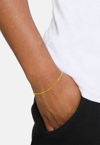 Bracciale in catena d'oro indossato su un polso, caratterizzato da un design attorcigliato e una superficie liscia. Abbinato a un indumento nero con tasche e una camicia bianca.