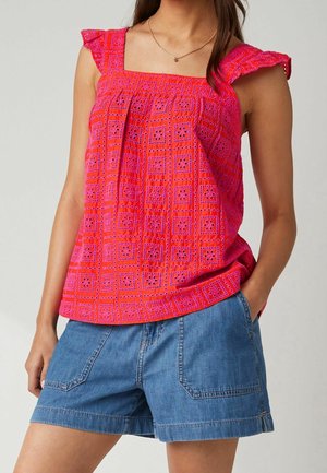 Kobieta w jaskraworóżowej, bezrękawnikowej bluzce w kwadratowy wzór oraz niebieskich dżinsowych szortach, z jedną ręką w kieszeni.