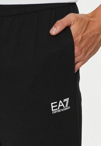 Pantaloni della tuta neri con logo "EA7" e "Emporio Armani" in bianco. Comprendono tasche laterali e una cintura elastica per il massimo comfort.