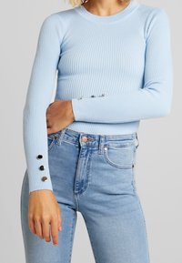 Kvinna som bär en ljusblå ribbad långärmad tröja med silverknappar på ärmarna och högmidjade ljusblå jeans.