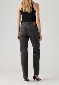 Mujer con largo cabello rubio de espaldas, vistiendo una blusa sin mangas negra, jeans de cintura alta gris oscuro y sandalias negras de tacón.