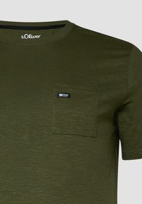 T-shirt vert olive à manches courtes avec col rond, poche poitrine avant gauche et petite étiquette noire indiquant "Elevated Casual."