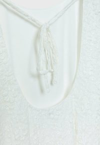 Haut sans manches blanc texturé avec broderie florale et fermeture à nouer tressée au niveau de l'encolure arrondie sur un fond clair.
