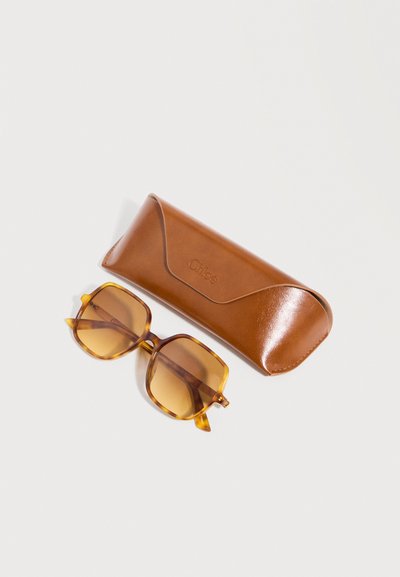 Lunettes de soleil carrées teintées marron avec montures en écaille de tortue ambrée, posées à côté d'un étui en cuir marron assorti sur un fond blanc.