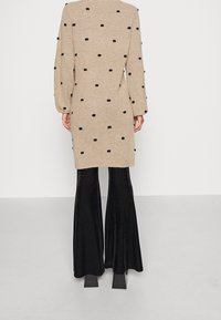 Tunique en maille beige à pois noirs, dotée de manches longues, d'une coupe décontractée et d'une fente haute sur les côtés, assortie à un pantalon noir évasé.