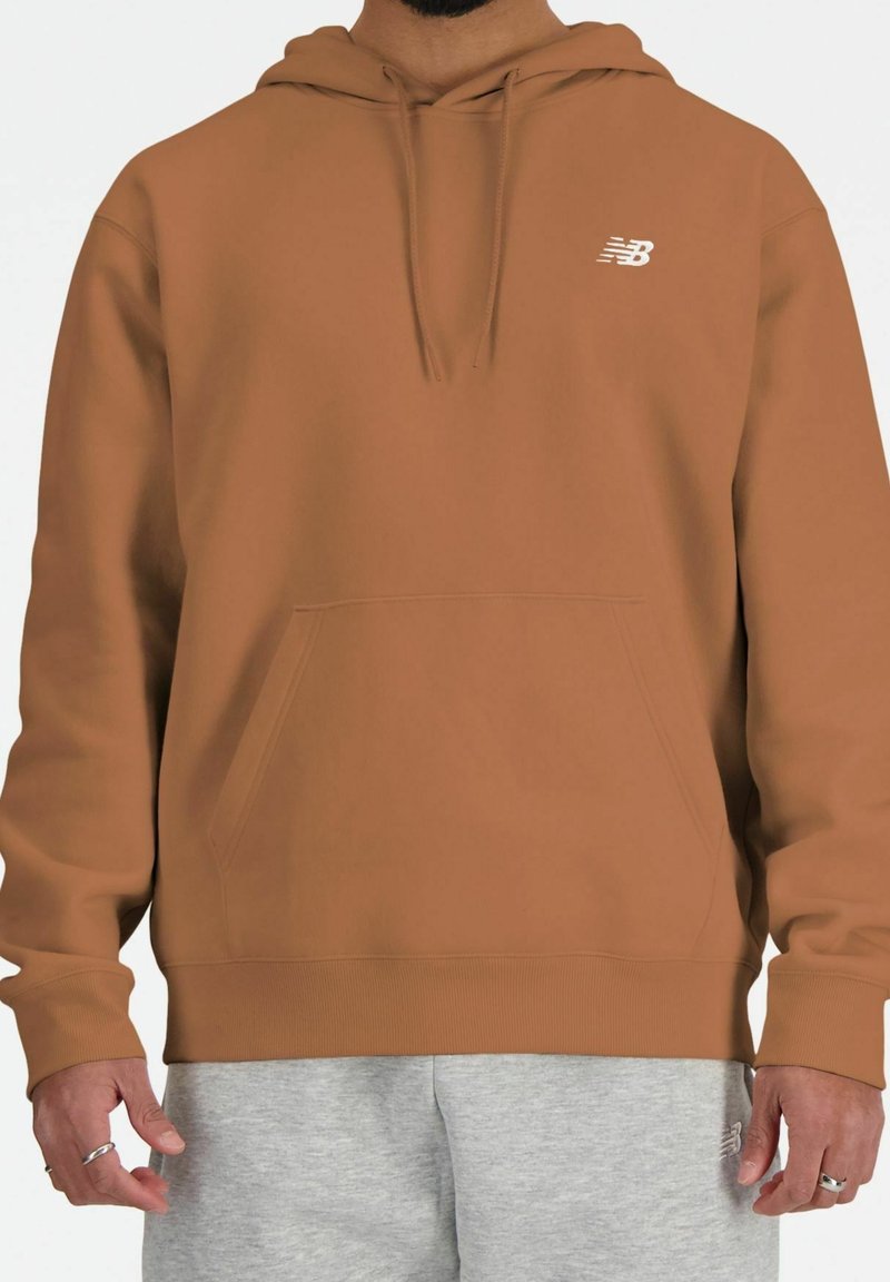 Homme portant un sweat à capuche New Balance marron et un pantalon de survêtement gris clair, debout avec les mains détendues le long du corps.