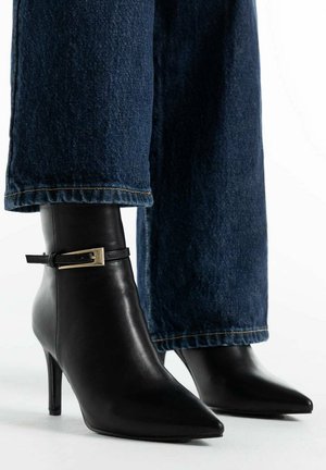 Stivaletti in pelle nera con punte appuntite e sottili cinghie con fibbia, indossati con pantaloni in denim blu scuro a gamba larga.
