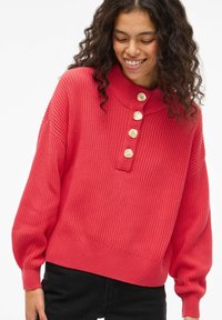 Roosa kootud kampsun kõrge kraega, soonikkoes tekstuuriga ja viie kuldse nupuga esiküljel. Omaduseks langevad õlad ja lühendatud serv.