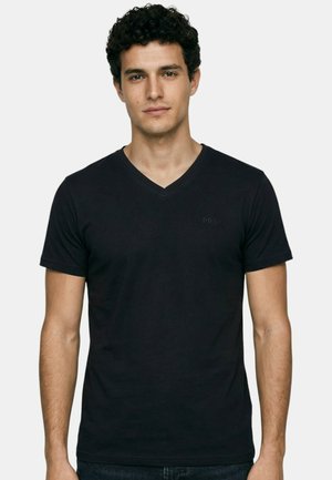 Jeune homme aux cheveux courts et bouclés portant un t-shirt noir uni à col en V, debout devant un fond blanc.
