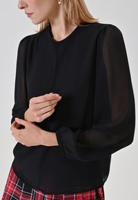 Schwarze Bluse mit durchsichtigen langen Ärmeln und weicher Textur. Mit runder Ausschnitt und dezenten Knopfdetails an der Vorderseite. Kombiniert mit einem karierten Rock.