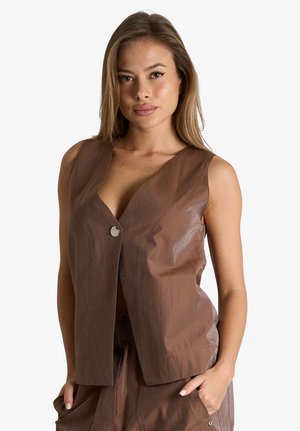 Giovane donna con capelli lunghi che indossa un gilet marrone senza maniche con bottoni e pantaloni coordinati, con una mano in tasca, sfondo neutro.