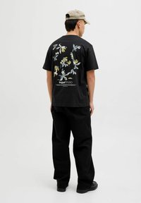 Schwarzes Baumwoll-T-Shirt mit floralem Druck in Pastellfarben auf dem Rücken, kombiniert mit lockeren schwarzen Hosen und schwarzem Schuhwerk.