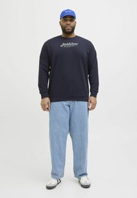 Marineblaues Sweatshirt mit bedrucktem Logo, blaue Kappe, hellblaue baggy Jeans und weiße Sneakers mit schwarzen Akzenten. Einfaches Design und lässige Passform.