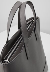 Bolso de mano gris con acabado texturizado, cierre con cremallera y asas dobles. Presenta un forro negro y un diseño minimalista.