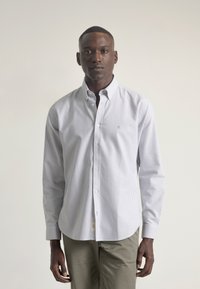 Camisa blanca de botones con rayas verticales en gris claro, de mangas largas, con cuello y un pequeño logo. Combinada con pantalones verdes oliva.