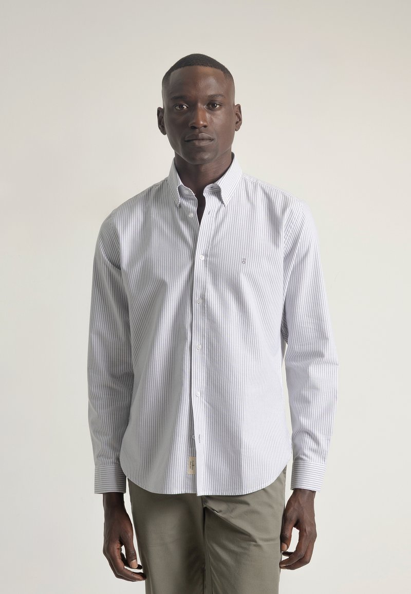 Camisa blanca de botones con rayas verticales en gris claro, de mangas largas, con cuello y un pequeño logo. Combinada con pantalones verdes oliva.