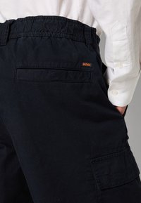 Gros plan sur un pantalon cargo noir avec une taille élastique et une étiquette "BOSS", porté avec une chemise blanche à manches longues, main dans la poche.