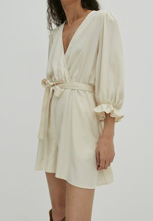 Femme portant une robe portefeuille couleur crème avec une taille nouée, un col en V et des manches bouffantes avec des poignets volantés, debout devant un fond uni.