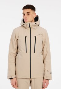 Beige waterdichte jas met capuchon, voorzien van zwarte ritsen, twee voorzakken en ventilatiegaten bij de kraag. Gladde textuur.