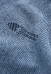 Jasnoniebieska tkanina o teksturowej powierzchni, z ciemnoniebieskim wytłoczonym logo z napisem "LOS ANGELES CALIFORNIA" z przodu.