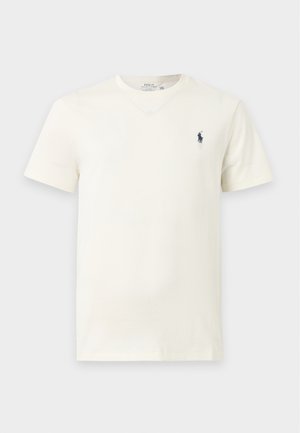 Νέος άνδρας που φοράει λευκό T-shirt, σκούρο παντελόνι, λευκά αθλητικά παπούτσια και καφέ μπερέ, στέκεται μπροστά σε ένα απλό φωτεινό φόντο.