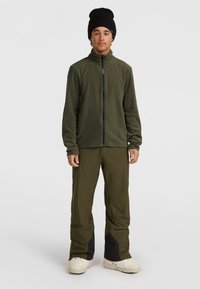 Grön fleece zip-jacka med hög krage, i kombination med gröna byxor med svarta accenter vid fållarna. Modellen bär en svart mössa och vita skor.