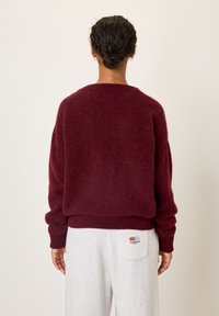 Personne aux cheveux bruns bouclés attachés en un petit chignon porte un pull bordeaux en tissu pelucheux et un pantalon gris clair avec un petit motif coloré sur la poche arrière.