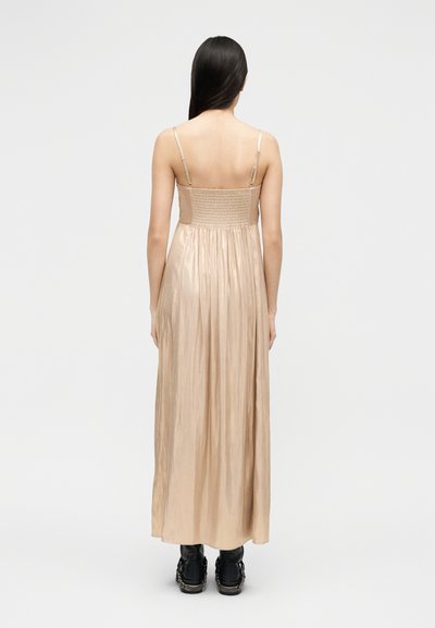 Γυναίκα με μακριά σκούρα μαλλιά φοράει λαμπερό μπεζ μακρύ φόρεμα χωρίς μανίκια με ρυθμιζόμενους ιμάντες, συνδυασμένο με μαύρες μπότες, στέκεται μπροστά σε απλό φόντο.