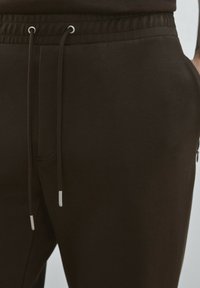 Braune Jogginghose aus weichem Material, mit elastischem Bund, Kordelzügen mit silbernen Enden und seitlichen Taschen.
