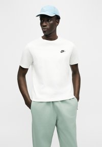 Νέος άνδρας που φοράει ανοιχτό μπλε καπέλο Nike, λευκό μπλουζάκι Nike και ανοιχτοπράσινο παντελόνι με τα χέρια στις τσέπες, στέκεται μπροστά σε απλό φόντο.