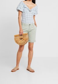 Modrý a bílý kostkovaný top s nafukovacími rukávy, zavazovaný vpředu, kombinovaný s světle zelenými šortkami a bambusovou kabelkou ve tvaru půlměsíce. Ploché sandály.