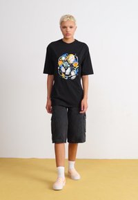 Black t-shirt with colorful graphic design, gray cargo shorts, white socks, and patterned shoes. Plain background and yellow floor.