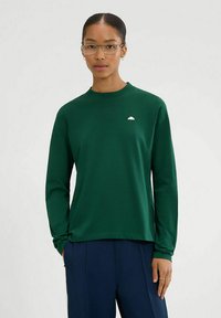 Top verde de manga larga hecho de tela suave con cuello redondo y pequeño logo blanco en el pecho, combinado con pantalones azul marino.