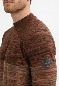 Moški nosi rjav in črn melirani pleten pulover z visokim ovratnikom in črnim Gabbiano etiketo na zgornjem delu rokava.