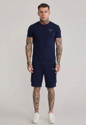 Mann mit umfangreichen Tattoos an Armen und Beinen trägt ein dunkelblaues SikSilk-T-Shirt und Shorts mit weißen Sneakers, stehend und nach vorne schauend.