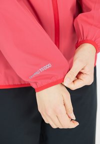 Röd vattentät jacka med elastiska ärmslut, har "W-PRO 15000"-logotyp på ärmen, slät textur och dragkedja fram.