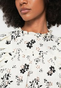 Λευκό φλοράλ μπλουζάκι με μαύρα και καφέ μοτίβα, λαιμόκοψη με βολάν, υφασμάτινη υφή και κοντά φουσκωτά μανίκια με λεπτομερές ντεκολτέ.