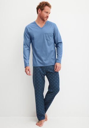 Mann trägt ein blaues, langärmeliges V-Ausschnitt-Shirt mit Brusttasche und marineblauen, gemusterten Schlafanzughosen, steht barfuß vor einem weißen Hintergrund.